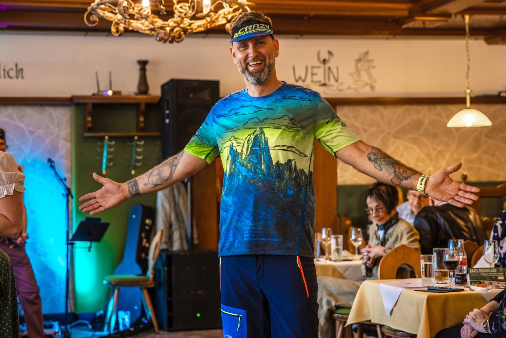 Ein Mann präsentiert sportliche Outdoorbekleidung auf dem Laufsteg einer Modenschau in einem Gasthaus. Er trägt ein bunt bedrucktes Funktionsshirt, dunkelblaue Shorts und ein sportliches Stirnband, während er mit ausgestreckten Armen lächelnd posiert. Die Gäste sitzen an den Tischen und verfolgen die Show in gemütlicher Atmosphäre.