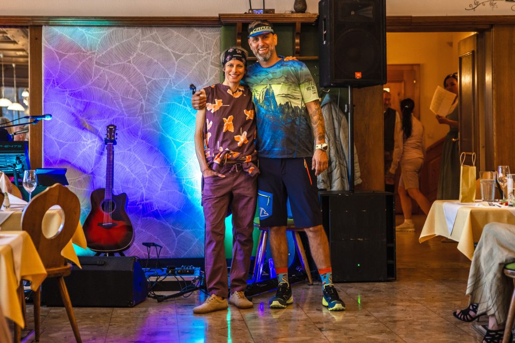 Zwei Personen posieren gemeinsam in farbenfroher Sport- und Outdoorbekleidung vor einer beleuchteten Bühne in einem Gasthaus. Die Frau trägt ein violettes Freizeitoutfit mit Stirnband, der Mann ein bunt bedrucktes Funktionsshirt mit Shorts, Cap und Sportschuhen. Eine Gitarre und stimmungsvolle Lichtakzente unterstreichen die lockere Atmosphäre der Modenschau.