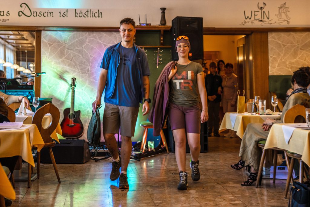 Zwei junge Erwachsene präsentieren funktionelle Sportmode auf einem Laufsteg in einem Gasthaus. Der Mann trägt ein blaues Outdoor-Shirt mit Shorts und Wanderschuhen, die Frau kombiniert ein sportliches Shirt mit kurzen Tights, Stirnband und Hiking-Boots. Im Hintergrund verfolgen Gäste die Modenschau bei stimmungsvoller Beleuchtung.
