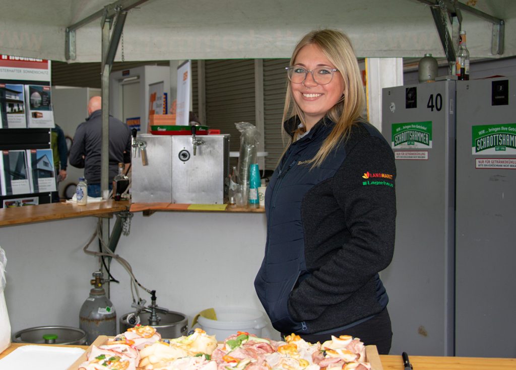 Eine Mitarbeiterin von Landmarkt Lagerhaus steht lächelnd am Bewirtungsbereich des Kompetenztags in Aigen. Im Vordergrund sind belegte Brötchen für die Gäste vorbereitet.
