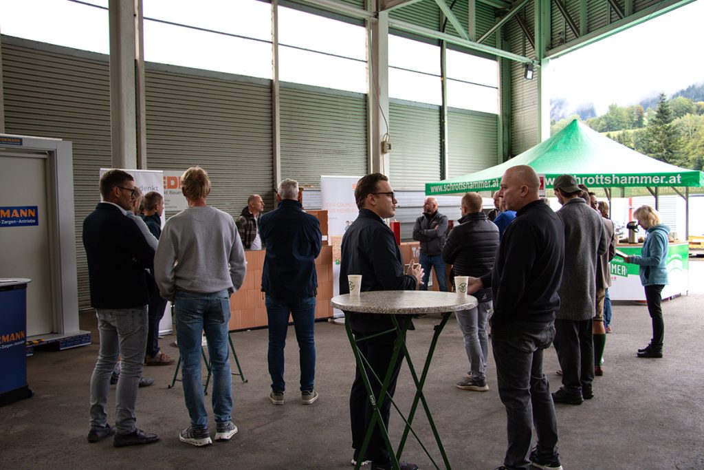 Mehrere Besucher stehen beim Kompetenztag in Aigen in kleinen Gruppen zusammen und tauschen sich vor verschiedenen Baustoff- und Partnerständen aus.