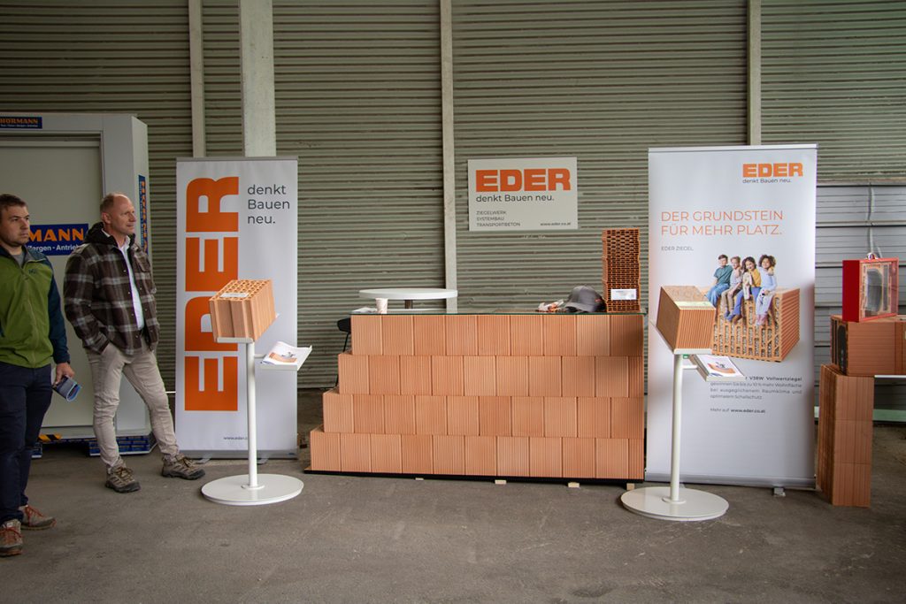 Ausstellungsbereich von Eder beim Kompetenztag in Aigen mit verschiedenen Ziegeln und Produktmustern. Zwei Besucher stehen neben dem Präsentationsstand und informieren sich über die gezeigten Baustofflösungen.