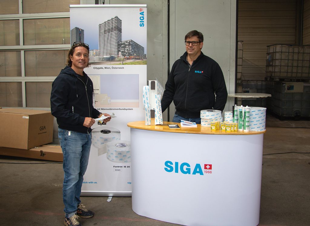 Zwei Fachberater stehen beim Kompetenztag in Aigen am SIGA-Informationsstand mit verschiedenen Produkten und Anschauungsmaterialien rund um Abdichtung und Gebäudehülle.