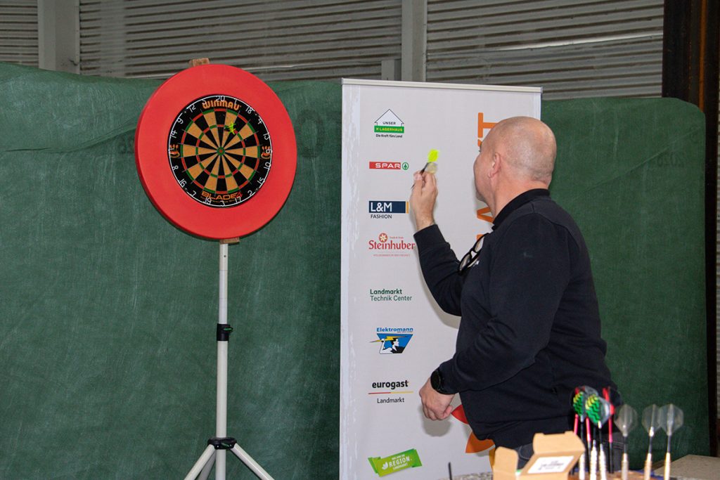 Ein Besucher nimmt beim Kompetenztag in Aigen an einer Dart-Challenge teil. Neben der Dartscheibe steht ein Landmarkt-Aufsteller mit den Logos verschiedener Unternehmensbereiche.