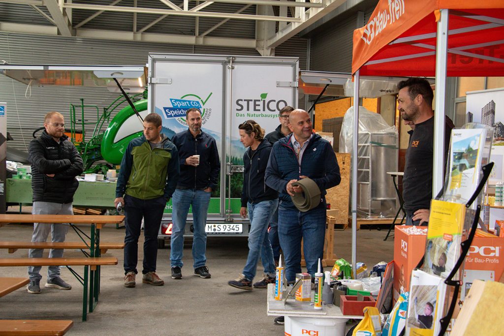 Mehrere Besucher stehen beim Kompetenztag in Aigen an den Informationsständen von PCI und Steico und tauschen sich mit Fachberatern über verschiedene Baustoffprodukte und Lösungen aus.
