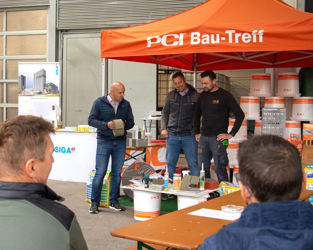 Drei Fachberater präsentieren beim Kompetenztag in Aigen verschiedene Bauprodukte am PCI Bau-Treff, während mehrere Besucher die Vorführung verfolgen.