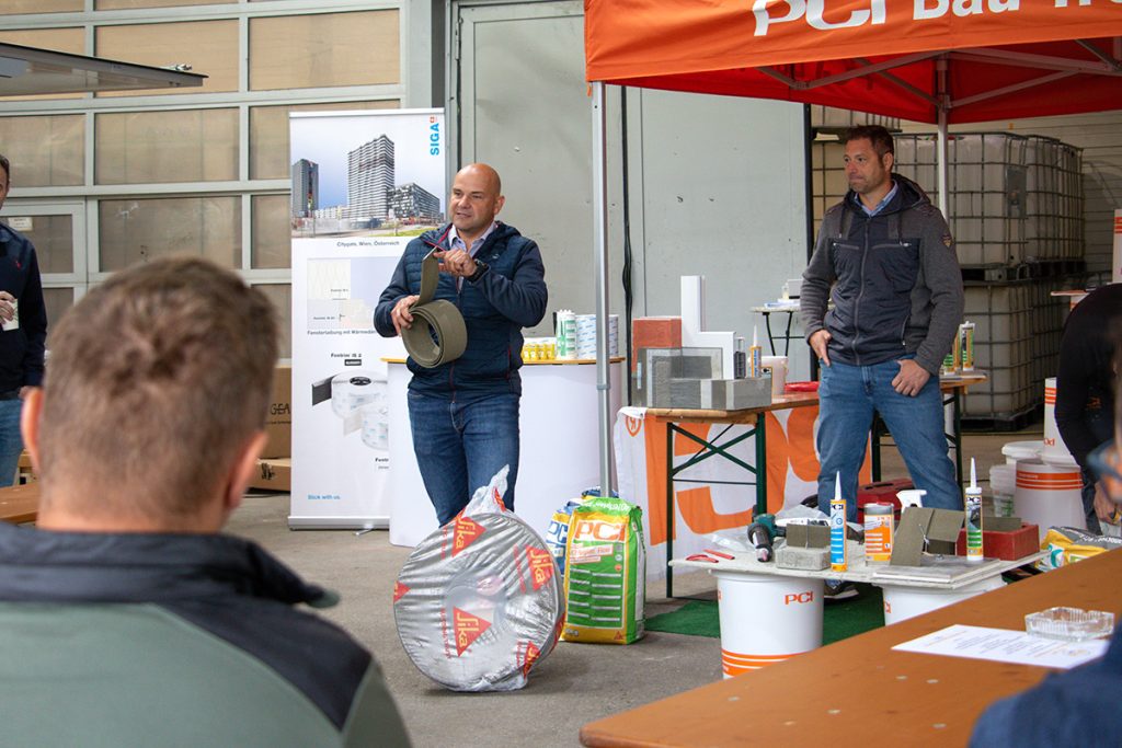 Ein Fachberater präsentiert beim Kompetenztag in Aigen ein Bauprodukt vor mehreren Besuchern. Im Hintergrund sind weitere Baustoffe und Informationsstände von PCI und SIGA zu sehen.