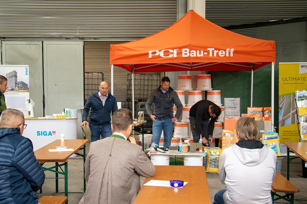 Mehrere Besucher verfolgen beim Kompetenztag in Aigen eine praktische PCI-Produktvorführung. Im Hintergrund sind verschiedene Baustoffprodukte sowie Informationsstände von PCI, SIGA und Isover zu sehen.