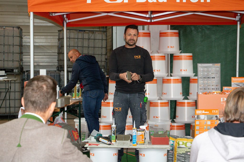 Ein PCI-Fachberater präsentiert beim Kompetenztag in Aigen verschiedene Baustoffprodukte und Anwendungslösungen vor mehreren Besucherinnen und Besuchern.