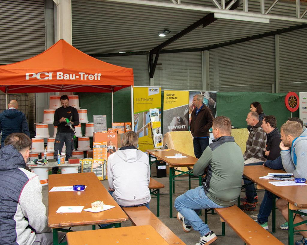 Mehrere Besucher verfolgen beim Kompetenztag in Aigen eine Produktvorführung am PCI Bau-Treff. Im Hintergrund sind unter anderem Informationsstände und Dämmstoffe von Isover zu sehen.