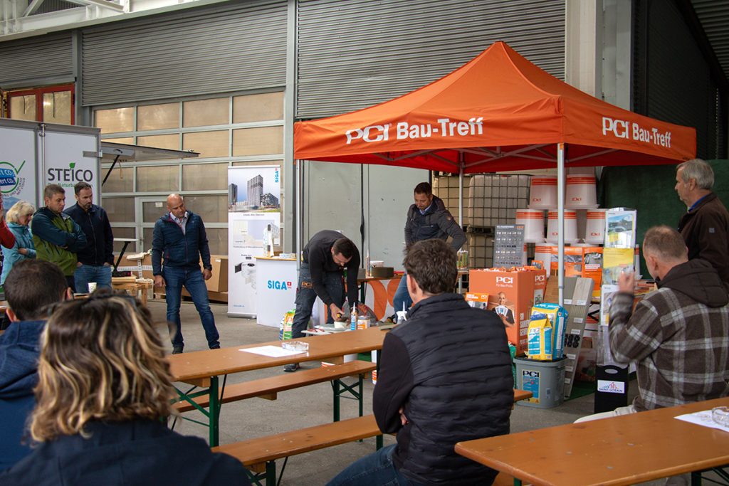 Mehrere Besucher verfolgen beim Kompetenztag in Aigen eine Fachvorführung am PCI Bau-Treff. Verschiedene Baustoffprodukte und Informationsstände sind rund um den Vorführbereich aufgebaut.