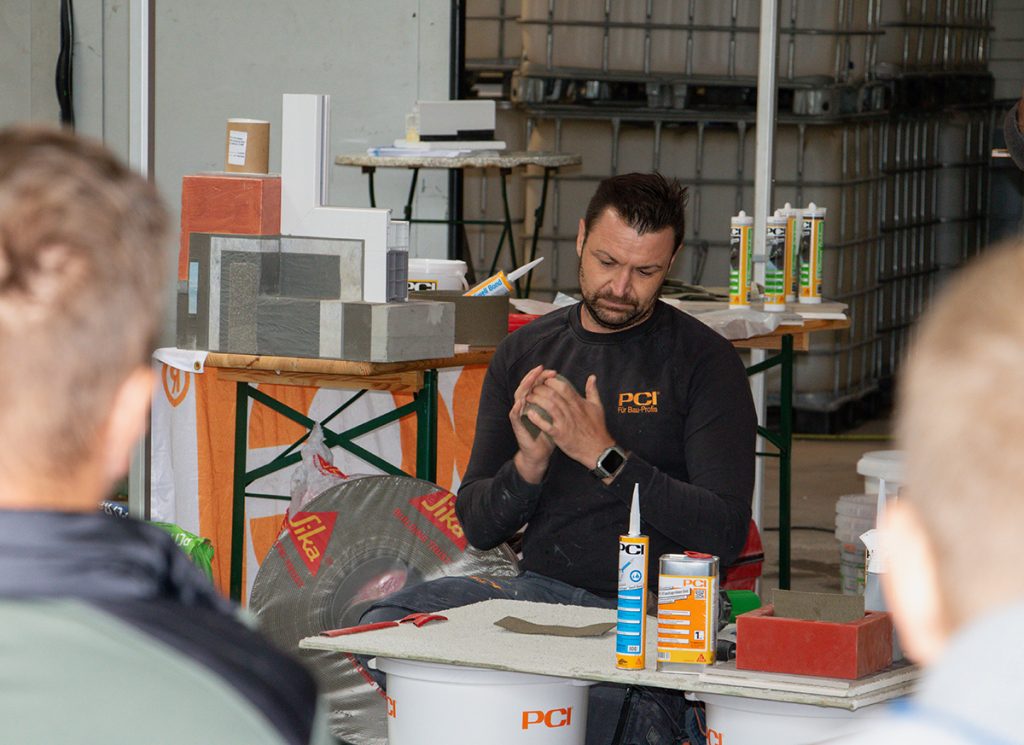 Ein PCI-Fachberater demonstriert beim Kompetenztag in Aigen vor mehreren Besuchern die Verarbeitung eines Baustoffprodukts. Verschiedene PCI-Produkte und Anschauungsmaterialien sind am Vorführtisch aufgebaut.